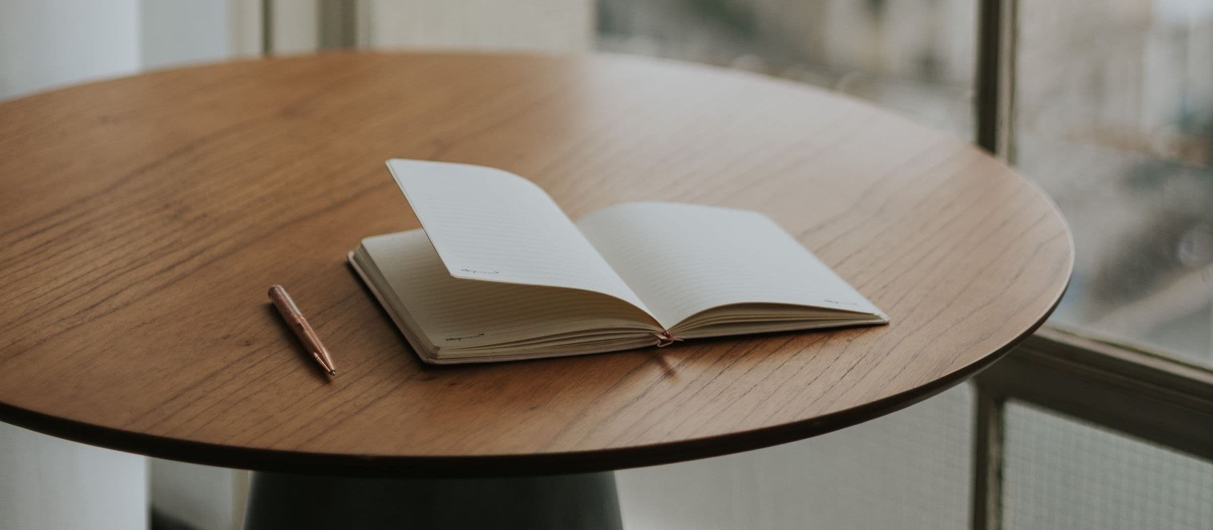 An open notebook and pen resting on a round wooden table by a window